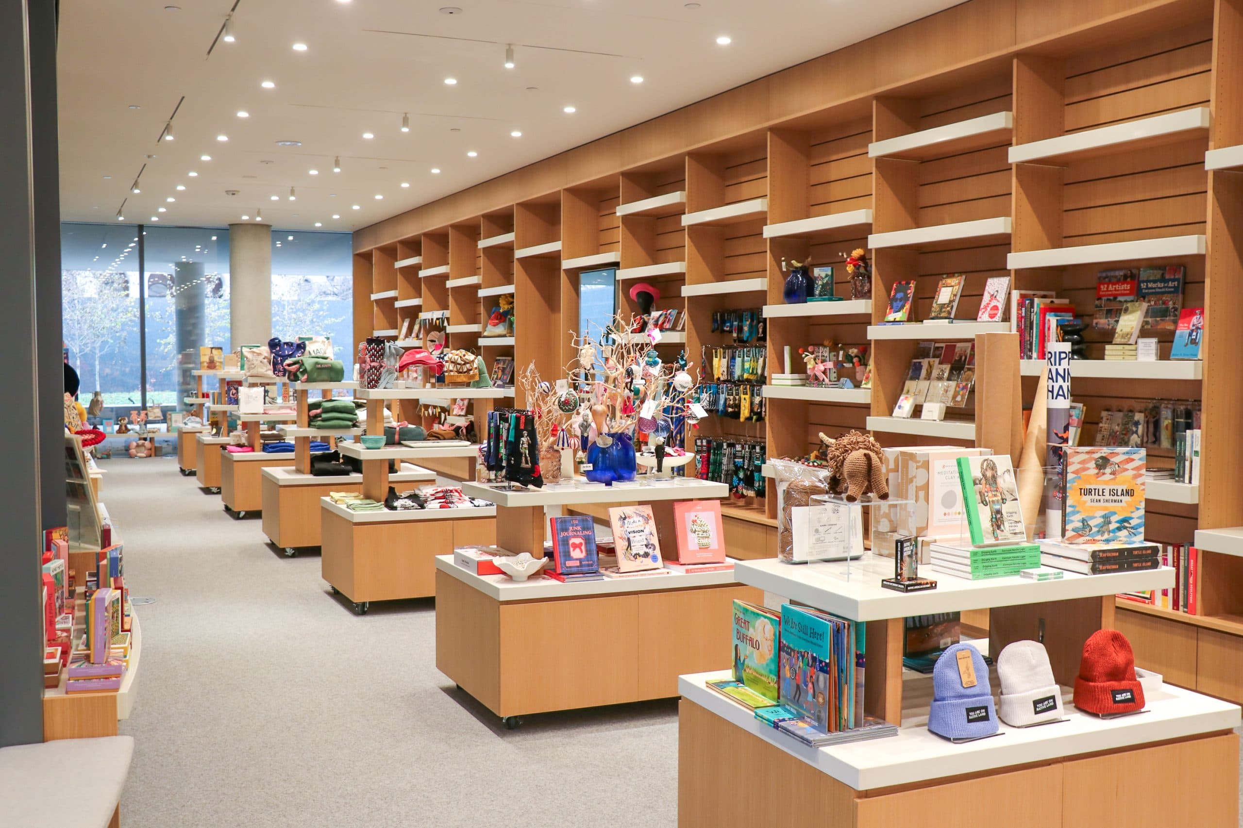 A modern bookstore with wooden shelves and display tables filled with books, toys, and various gift items. The space is well-lit, spacious, and organized, with large windows at the back.