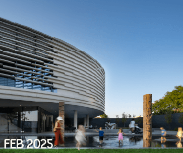 Children play in a shallow water feature near a modern, curved building with layered white exterior panels under a clear blue sky. The date "FEB 2025" appears in the bottom left corner.