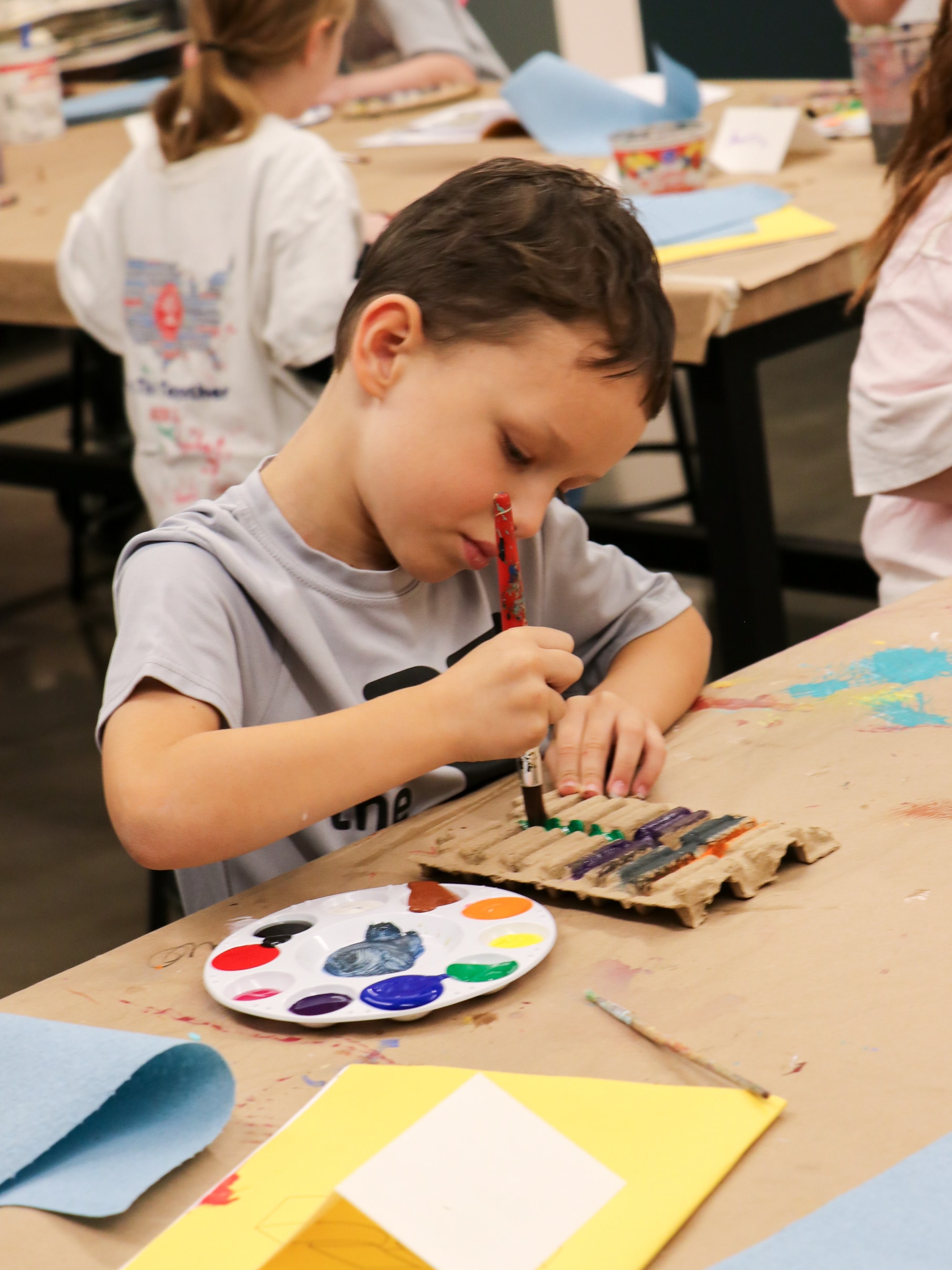 Young child concentrating on painting a small cardboard texture with a paintbrush, surrounded by art supplies and wearing a light shirt. Other children are partially visible in the background, engaged in similar activities.