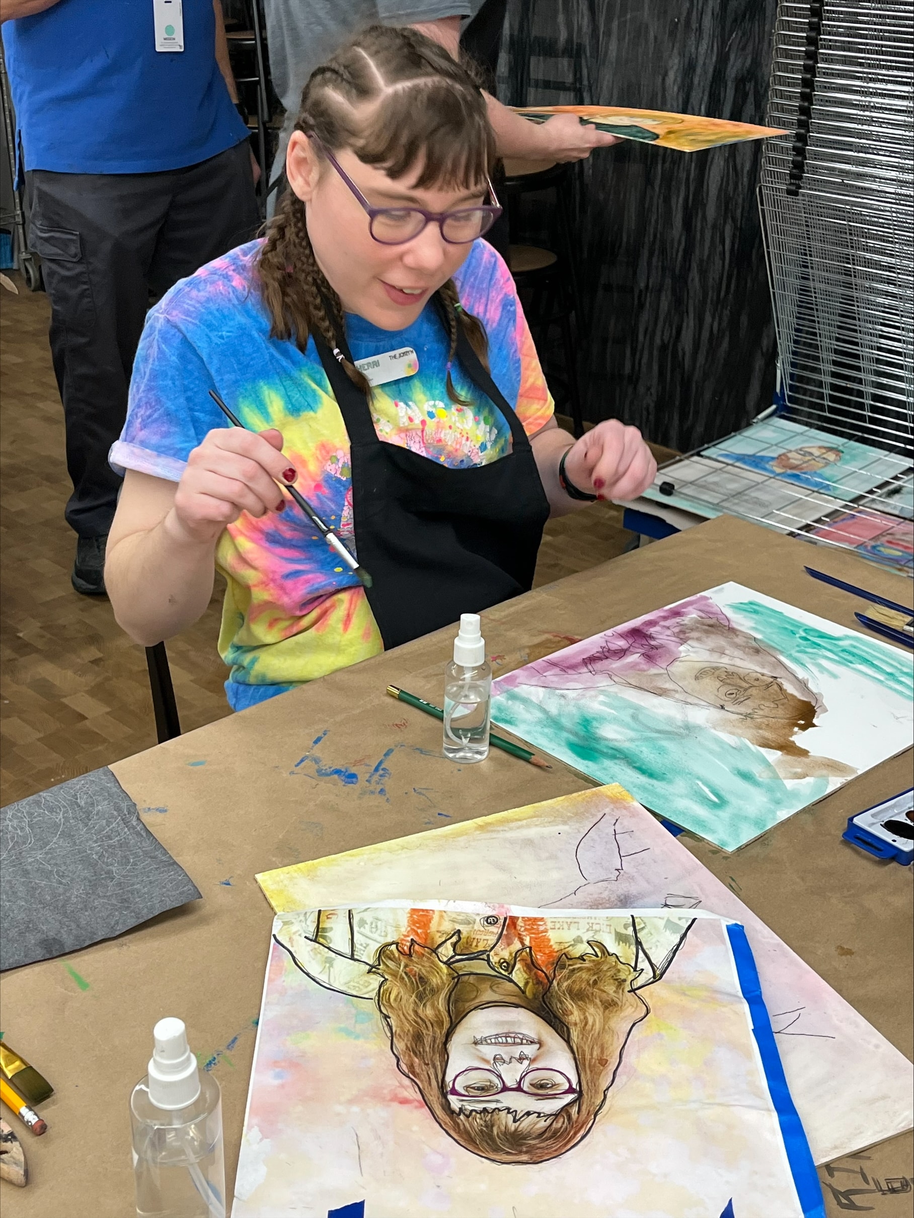 A person wearing glasses, a tie-dye shirt, and an apron is painting at a table. In front of them is a watercolor portrait on paper. Art supplies are scattered on the table, and they are holding a brush, smiling and engaged in the activity.